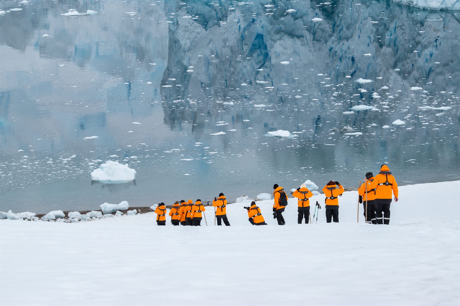 Line of visitors in orange parkas walking across snow beside a glacier