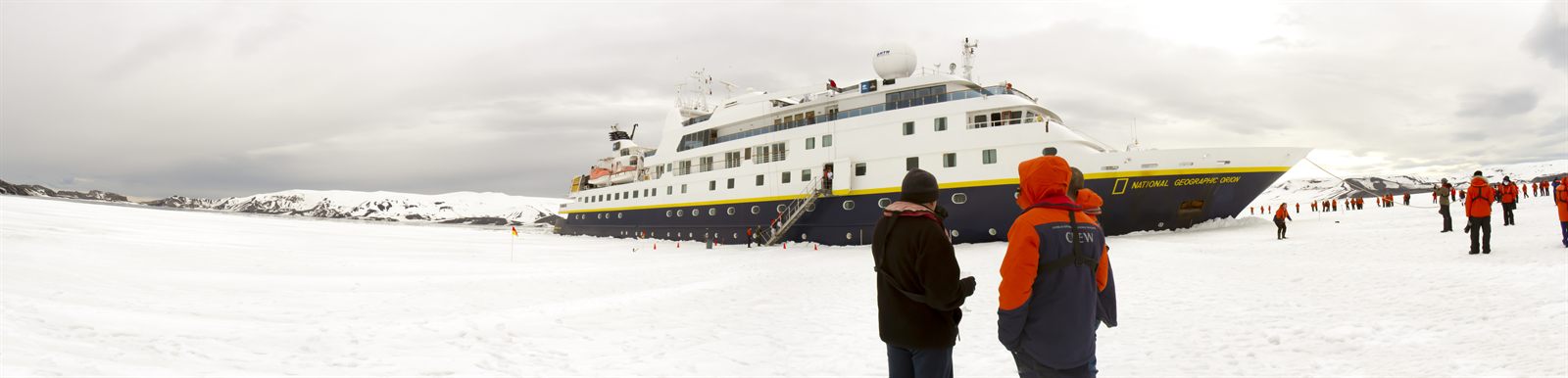 Expedition ship anchored off a snowfield while passengers in red parkas walk ashore