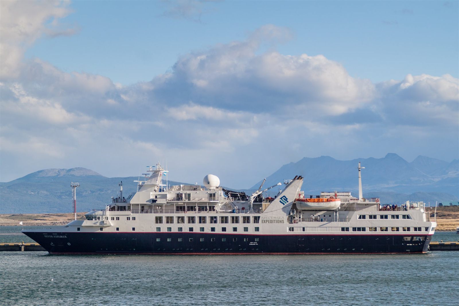 Small blue-hulled expedition ship alongside a pier below mountains